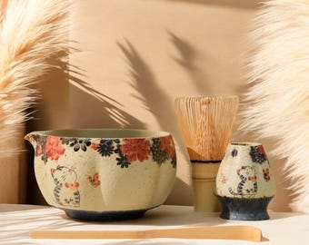 Op de afbeelding: Een keramische matcha-theeset met een kom, een klopper, een klopperhouder en een bamboelepel. De kom en houder zijn versierd met bloemen- en kattenmotieven in rood, zwart en blauw. De set staat op een wit oppervlak.