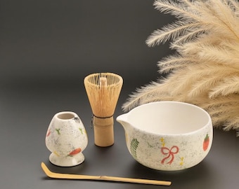 Könnte beinhalten: Ein Matcha-Teeset mit einem Bambusbesen, einer Keramikschale mit Ausguss, einem kleinen Keramikhalter und einem Bambuslöffel. Schale und Halter haben ein gesprenkeltes Design mit roten und grünen Akzenten.