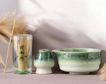May include: A matcha tea set featuring a bamboo whisk in a clear container with Japanese text, a small ceramic tea bowl, and a larger green and white ceramic bowl with a spout. The set is arranged on a neutral surface.
