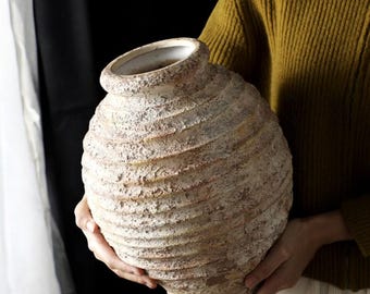 May include: A large, textured, beige ceramic vase with a wide, spiraling design. The vase has a rounded body and a slightly flared rim. The surface has a rough, aged appearance, suggesting a rustic or antique style. The vase is held by a person.