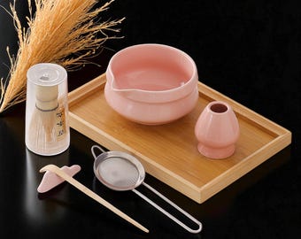 Puede incluir: Un juego de té matcha con un cuenco de cerámica rosa, un soporte para batidor y un batidor de bambú en una funda transparente. El juego incluye un colador de acero inoxidable, una cuchara de bambú y una bandeja de madera. Los artículos están sobre una superficie negra.