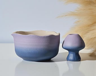 Può includere: Una ciotola in ceramica e un vaso a forma di fungo. La ciotola ha un bordo ondulato e una sfumatura da blu a viola. Il vaso a forma di fungo è anch'esso blu-viola. Entrambi sono su una superficie bianca con uno sfondo azzurro.