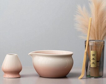 May include: A matcha tea set with a light pink and white color scheme. The set includes a ceramic bowl, a whisk, and a whisk holder. A bamboo whisk and a container with Japanese text are also present.