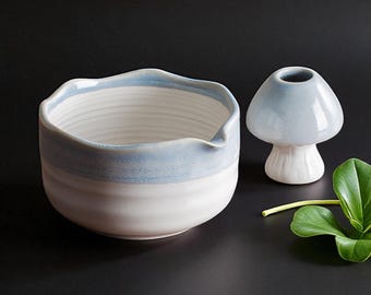 Puede incluir: Un cuenco de cerámica con borde azul claro y cuerpo blanco, junto a un jarrón con forma de hongo. El cuenco tiene un borde ondulado y un pico. El jarrón es azul claro en la parte superior y blanco en la base. Una hoja verde está en primer plano.