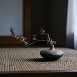 Op de afbeelding: Een kleine, donkergrijze keramische vaas met een afgeronde body en een smalle hals. De vaas bevat een delicaat, gedroogd bloemstuk met dunne, bruine stelen en kleine, getextureerde bloesems. De vaas staat op een getextureerd, neutraal getint oppervlak.