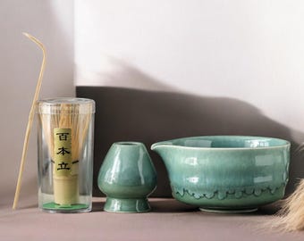 May include: A matcha tea set featuring a green ceramic bowl, a small green ceramic whisk holder, and a clear container with a bamboo whisk. A bamboo scoop rests beside the container. The set is arranged on a neutral surface.