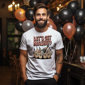 May include: White t-shirt with the text "LET'S GET MASHED" in a retro font. The shirt features a graphic of cartoon potatoes celebrating with champagne. The background includes black, orange, and gray balloons.