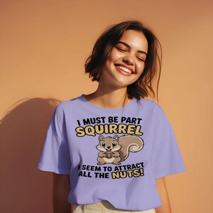 May include: A light purple t-shirt with the text "I MUST BE PART SQUIRREL" and "I SEEM TO ATTRACT ALL THE NUTS!" A cartoon squirrel holding a nut is in the center. The person wearing the shirt is smiling.