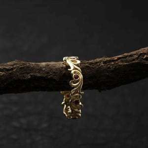 May include: A gold ring featuring an elaborate, swirling pattern and small red gemstones. The ring is presented on a dark, textured branch against a dark backdrop. The ring's intricate details are highlighted, showcasing its craftsmanship and design.