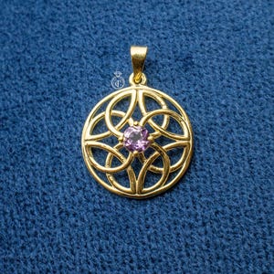 May include: A gold pendant featuring a Celtic knot design with a central purple gemstone. The pendant has a small loop at the top. The background is a textured blue fabric.