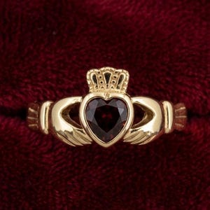 Könnte beinhalten: Ein goldfarbener Claddagh-Ring mit einem herzförmigen, dunkelroten Edelstein. Der Ring hat eine Krone über dem Herzen und zwei Hände, die das Herz umfassen. Der Ring ist vor einem dunkelroten Samthintergrund platziert.