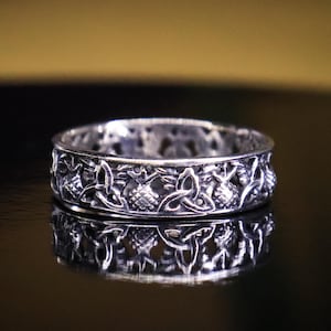 May include: A silver ring with an intricate Celtic knot design. The ring is highly detailed, featuring interwoven patterns and a reflective surface. The ring is displayed on a dark, reflective surface, highlighting its design.