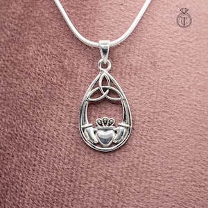 May include: A silver-toned pendant necklace featuring a teardrop-shaped design. The pendant showcases a Claddagh symbol with a heart, crown, and hands, and a Celtic knot at the top. The necklace is on a pink background.
