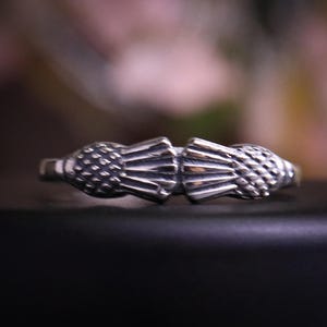 Op de afbeelding: Een zilveren ring met een gedetailleerd ontwerp van twee gestileerde ananasvormen die tegenover elkaar staan. De ring heeft een getextureerd oppervlak en staat tegen een vage achtergrond, wat het ingewikkelde metaalwerk benadrukt. De ring is een sieraad.