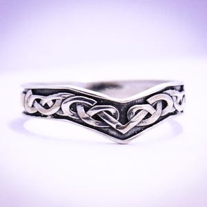 Op de afbeelding: Een zilverkleurige ring met een Keltisch knoopontwerp. De ring heeft een V-vormige band met ingewikkelde, verweven patronen. Het metaal heeft een licht geoxideerde uitstraling, wat de details van de knopen benadrukt. De achtergrond is een zachte gradiënt van wit en paars.