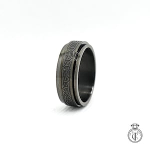 Peut inclure: Une bague gris foncé avec un motif de nœud celtique. La bague a une bande centrale rotative et un motif détaillé sur les bords extérieurs. La bague est posée sur un fond blanc.