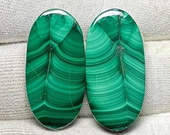 May include: Two oval-shaped malachite gemstones. The polished stones display a vibrant green color with intricate swirling patterns and bands. The gemstones are set against a neutral background.
