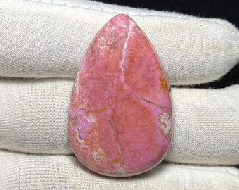 May include: A teardrop-shaped gemstone with a pink and reddish-brown color palette. The stone has a polished surface with visible veining and variations in color. The gemstone is held against a white fabric background.