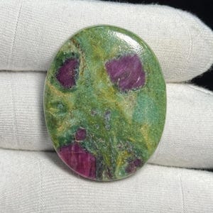 Könnte beinhalten: Ovaler Edelstein mit grüner Grundfarbe und leuchtend magentafarbenen Einschlüssen. Der polierte Stein hat eine glatte, glänzende Oberfläche, die die natürlichen Muster und Farben des Minerals hervorhebt. Der Edelstein wird in weißem Stoff gehalten.