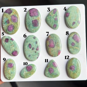 May include: Twelve polished cabochon gemstones arranged on a white display board, numbered one through twelve. Each stone features a light green base with embedded magenta-colored inclusions, displaying varied oval and triangular shapes.