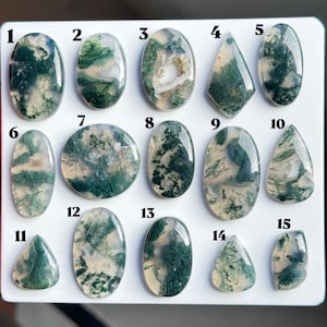 Puede incluir: Una colección de quince cabujones de ágata musgosa pulidos dispuestos sobre un tablero blanco. Cada piedra preciosa presenta inclusiones dendríticas verdes únicas que recuerdan al musgo o helechos en una base de cuarzo blanco lechoso, numeradas del 1 al 15.