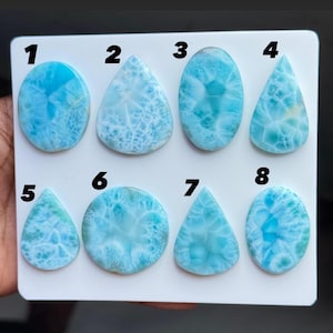 May include: Eight polished Larimar cabochons arranged on a white display board, numbered one through eight. Each stone features a distinct sky-blue and white marbled pattern with varying shapes, including ovals and teardrops, showcasing natural textures.