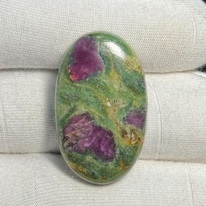 Puede incluir: Gema de forma ovalada con superficie pulida. La piedra presenta una vibrante mezcla de colores, incluyendo magenta intenso y verde. Las áreas magenta están agrupadas, creando un contraste llamativo sobre el fondo verde. Una piedra natural única.