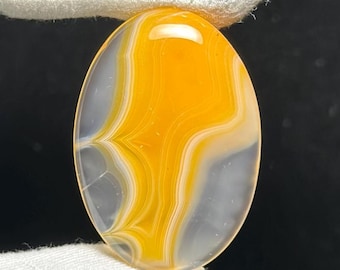 May include: Oval-shaped gemstone with a polished surface. The stone features a vibrant yellow and white banded pattern, with areas of translucent gray. The gemstone is held between two white fabric surfaces.