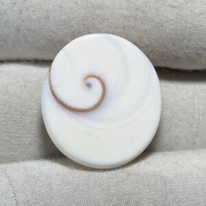 Pode incluir: Um pingente redondo de concha branca com um design em espiral em tons de castanho. O pingente tem uma superfície lisa e polida e provavelmente é feito de material natural. O detalhe em espiral adiciona um toque único e orgânico.