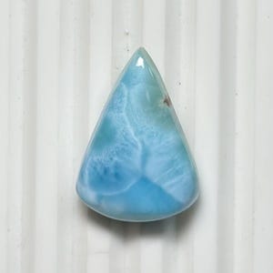 May include: A polished, teardrop-shaped gemstone with a light blue hue, featuring swirling white patterns. The stone has a smooth, glossy surface and is set against a white background. This gemstone is likely a Larimar.