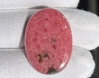 May include: Oval-shaped, polished gemstone in shades of pink and red, with dark brown and black inclusions. The stone is held in a white gloved hand, showcasing its smooth, reflective surface. The gemstone's color and pattern are the focal point.