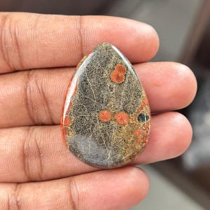 May include: A teardrop-shaped gemstone with a polished surface. The stone features a network of grey and brown patterns, with scattered orange-red circular inclusions. The gemstone is held in a hand.