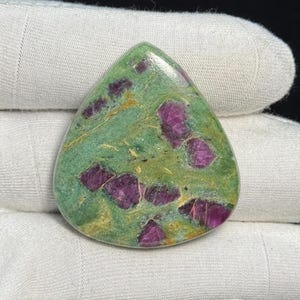 Puede incluir: Una gema en forma de lágrima con una base verde y parches irregulares de magenta intenso. La piedra pulida tiene una superficie brillante, con inclusiones de magenta dispersas por toda la piedra.