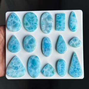 Puede incluir: Una colecci&oacute;n de catorce cabujones de larimar pulidos dispuestos sobre un tablero blanco. Las piedras presentan varias formas, incluyendo gotas, &oacute;valos, rect&aacute;ngulos y tri&aacute;ngulos, todas con un distintivo patr&oacute;n jaspeado azul claro.