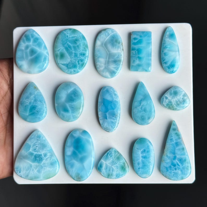 Puede incluir: Una colecci&oacute;n de catorce cabujones de larimar pulidos dispuestos sobre un tablero blanco. Las piedras presentan varias formas, incluyendo gotas, &oacute;valos, rect&aacute;ngulos y tri&aacute;ngulos, todas con un distintivo patr&oacute;n jaspeado azul claro.