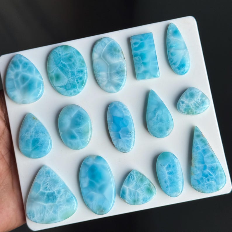 Puede incluir: Una colecci&oacute;n de catorce cabujones de larimar pulidos en varias formas, incluyendo &oacute;valos, gotas y un rect&aacute;ngulo. Cada piedra muestra un color azul cielo distintivo con patrones blancos similares a telara&ntilde;as y superficies lisas y brillantes.