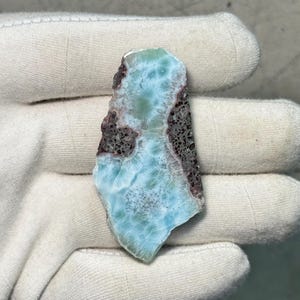 May include: A polished, irregular-shaped gemstone with a light blue and white pattern, reminiscent of ocean waves. The edges are a dark brown color. The stone is held in a white glove.