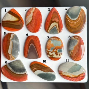 May include: Eleven polished, multi-coloured cabochon gemstones arranged on a white board, numbered one through eleven. Each stone features unique swirling patterns of orange, red, brown, grey, and cream, with smooth, rounded surfaces.