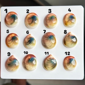 May include: A white display card featuring twelve cabochons arranged in three rows of four. Each cabochon has a smooth, glossy, domed surface with a swirling pattern of tan, brown, and blue colours. The items are numbered one through twelve.