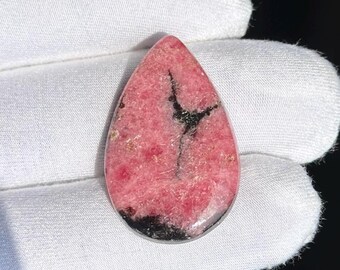 Könnte beinhalten: Ein tropfenförmiger Edelstein mit einem rosa und schwarzen Muster. Der polierte Stein hat einen leuchtenden rosa Farbton mit dunklen schwarzen Adern und Flecken. Der Edelstein wird vor einem weißen Hintergrund gehalten.