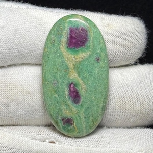 May include: An oval gemstone featuring a green base with striking, irregular patches of deep red. The polished stone displays red inclusions throughout, forming a unique pattern. The gemstone is set against a white background, highlighting its colours.