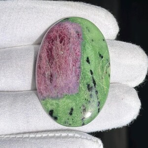 Puede incluir: Gema de forma ovalada con una vibrante combinación de colores. La piedra presenta una zona prominente de tonos rosa intenso y púrpura, contrastada por un campo circundante de color verde con motas negras. La superficie pulida refleja la luz.