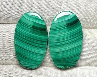 May include: Two polished oval malachite gemstones. The gemstones display a vibrant green color with distinctive banded patterns. The stones are set against a neutral, textured background, highlighting their natural beauty and intricate designs.