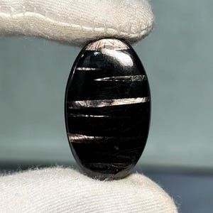 Puede incluir: Una piedra preciosa negra pulida de forma ovalada, con bandas blancas horizontales iridiscentes. La piedra se sostiene entre dos dedos cubiertos de tela blanca, mostrando su superficie lisa y reflectante. El color oscuro de la piedra contrasta con las bandas blancas.
