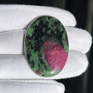 Op de afbeelding: Ovale robijn-zoisiet edelsteen. De steen heeft een levendige mix van kleuren, waaronder diep rood, zwart en groen. Het gepolijste oppervlak van de steen reflecteert licht en benadrukt de natuurlijke patronen en texturen.