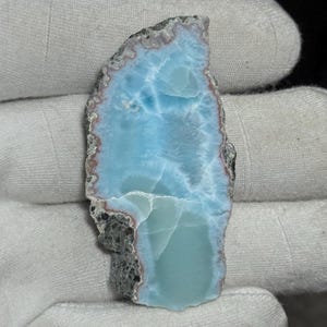 Puede incluir: Una gema pulida de forma irregular, con un patrón azul claro y blanco que recuerda a las olas del océano. Los bordes son una mezcla de gris y marrón, con una textura natural rugosa. La piedra se sostiene en un guante blanco.