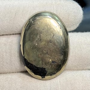 Pode incluir: Pedra preciosa de pirite em forma oval com um brilho metálico dourado. A superfície tem uma aparência rachada e texturizada com inclusões escuras e pretas. A pedra preciosa é segurada em uma luva branca, destacando sua beleza natural. Um espécime mineral polido único.