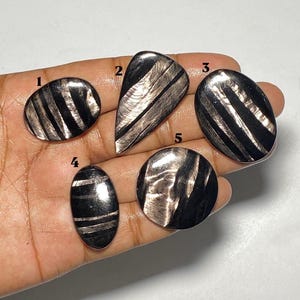 May include: Five polished cabochons with a black and metallic silver striped pattern, labeled with numbers 1 through 5. The stones feature smooth, reflective surfaces with distinct dark bands alternating with shimmering, light-reflecting metallic streaks.