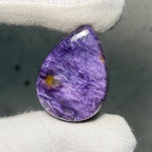Puede incluir: Una gema pulida en forma de lágrima, con un patrón arremolinado y fibroso en tonos morados y toques de amarillo. La piedra está sujeta por una tela blanca, mostrando su superficie lisa y reflectante.