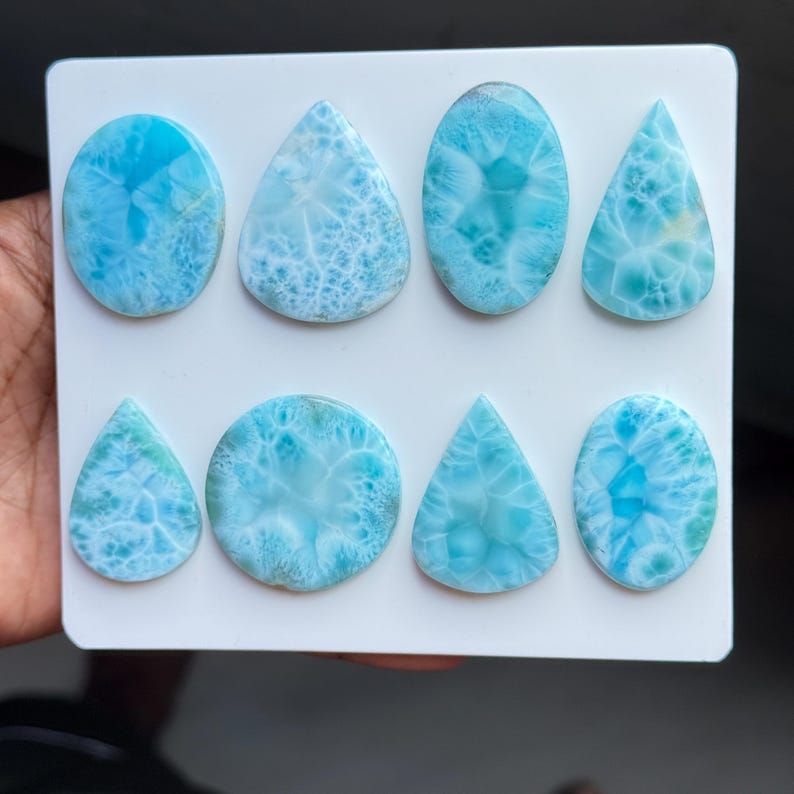 May include: Eight polished Larimar gemstones arranged on a white rectangular display card. The stones feature varying shades of sky blue and white with distinct web-like patterns, presented in a mix of oval, round, and teardrop shapes.